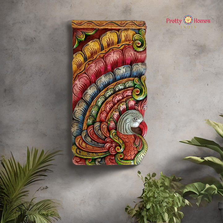 A multicolored woodcarved peacock wall hanging with fine detailing, displayed against a wall with plants around it.