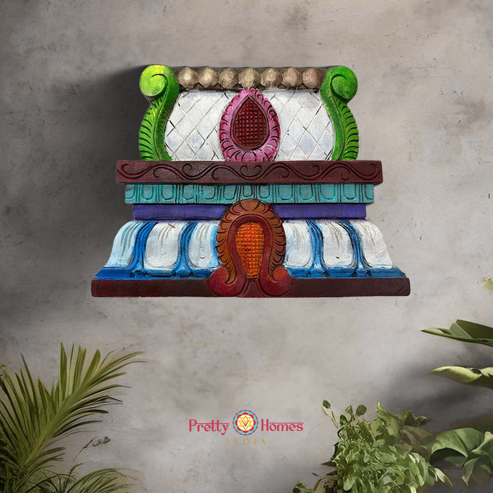 A multicolored, woodcarved temple mandir top with a dome design, featuring intricate carvings and a vibrant finish, displayed on a wall with a green and pink floral hanging in the background.
