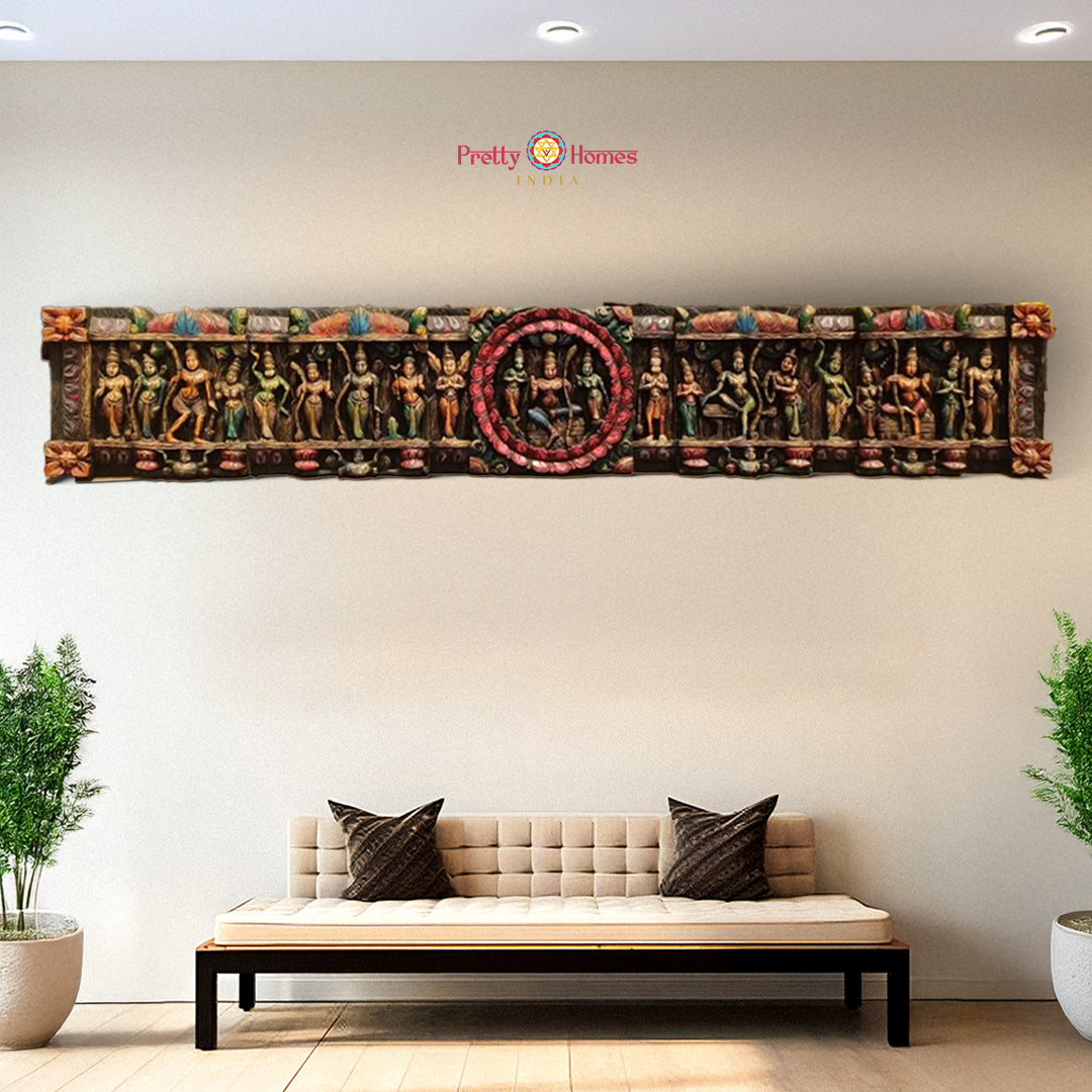 A horizontal wooden wall panel featuring a series of carved characters from the Hindu epic Ramayana, including Lord Ram, Hanuman, and Laxman, with a halo circle in the center. The panel is multicolored and placed above a modern day bench and potted plants in a room.