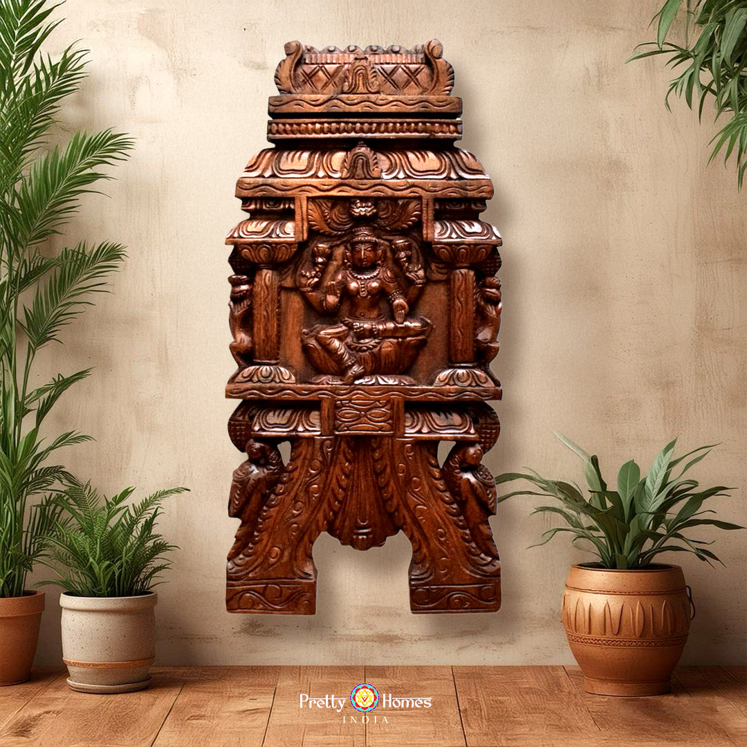 A wall-mounted wooden sculpture of a Hindu goddess Laxmi with two peacocks, surrounded by intricate carvings, in a room with potted plants and a decorative ceiling.