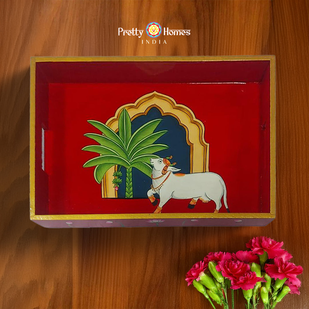 Red Pichwai wooden serving tray with hand-painted cow under palm tree and arched window.