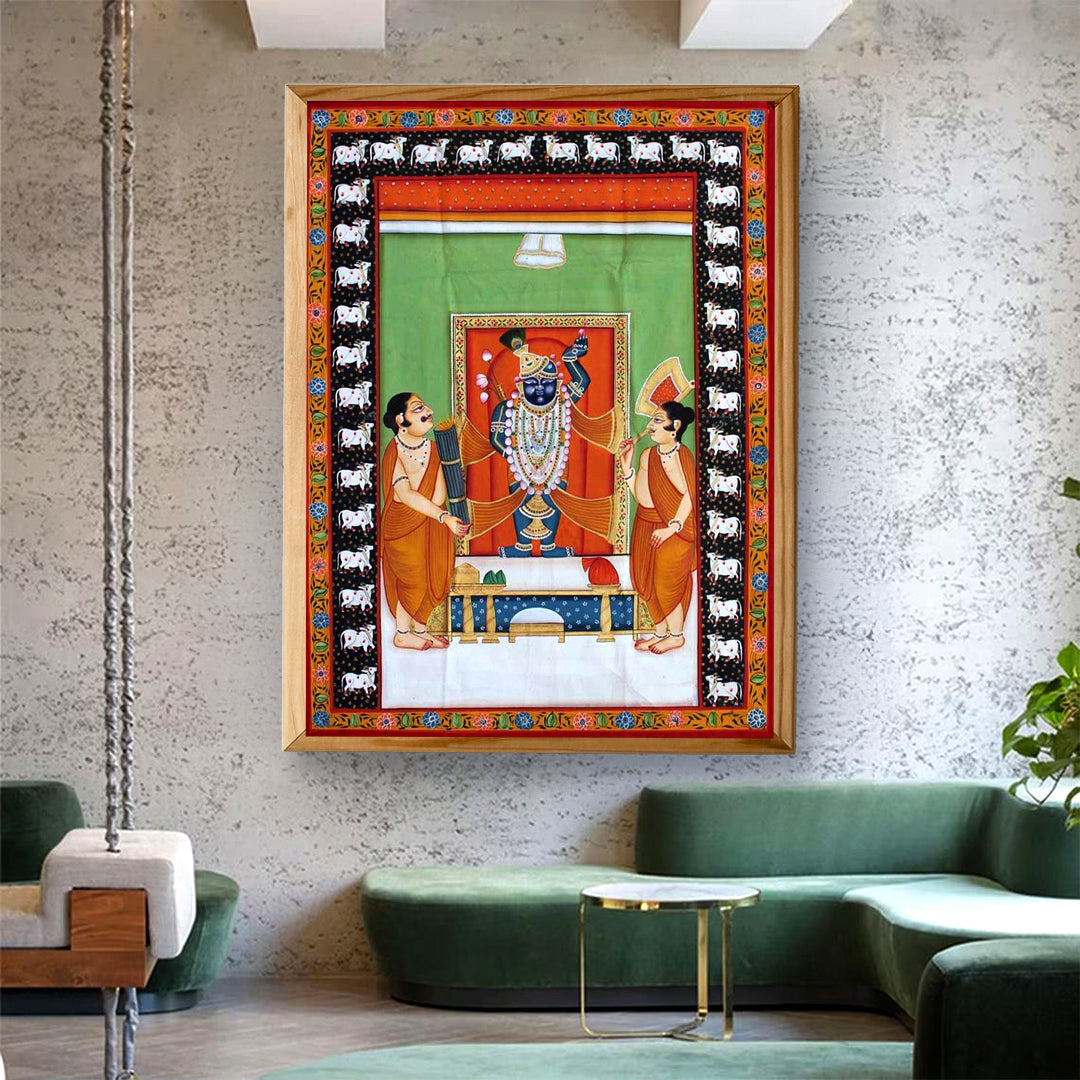 Framed Pichwai Shrinathji painting with two attendants and cow border, displayed in a styled living room.