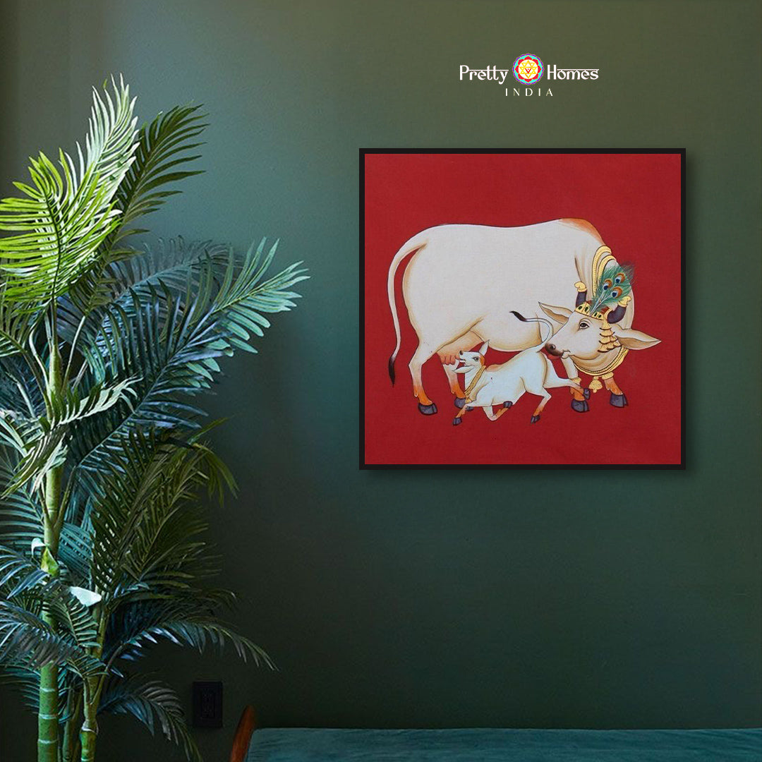Hand-painted pichwai featuring Kamadhenu and calf on red background, displayed in a serene home setting.