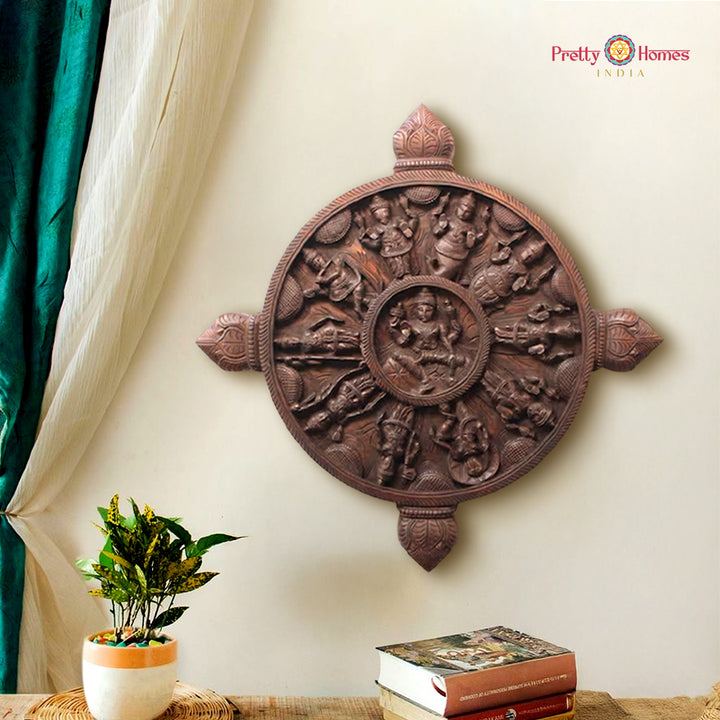 Decorative wooden wall panel with intricate carvings on a beige wall, featuring a plant and books on a table. Round carved wooden wall panel with Dasavatara and Ashtalakshmi center.

