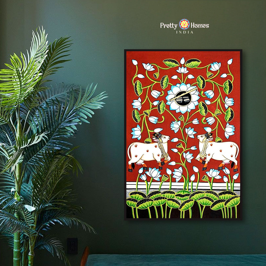 Hand-painted pichwai of two white cows standing among blooming lotuses on red background, displayed in a modern room.