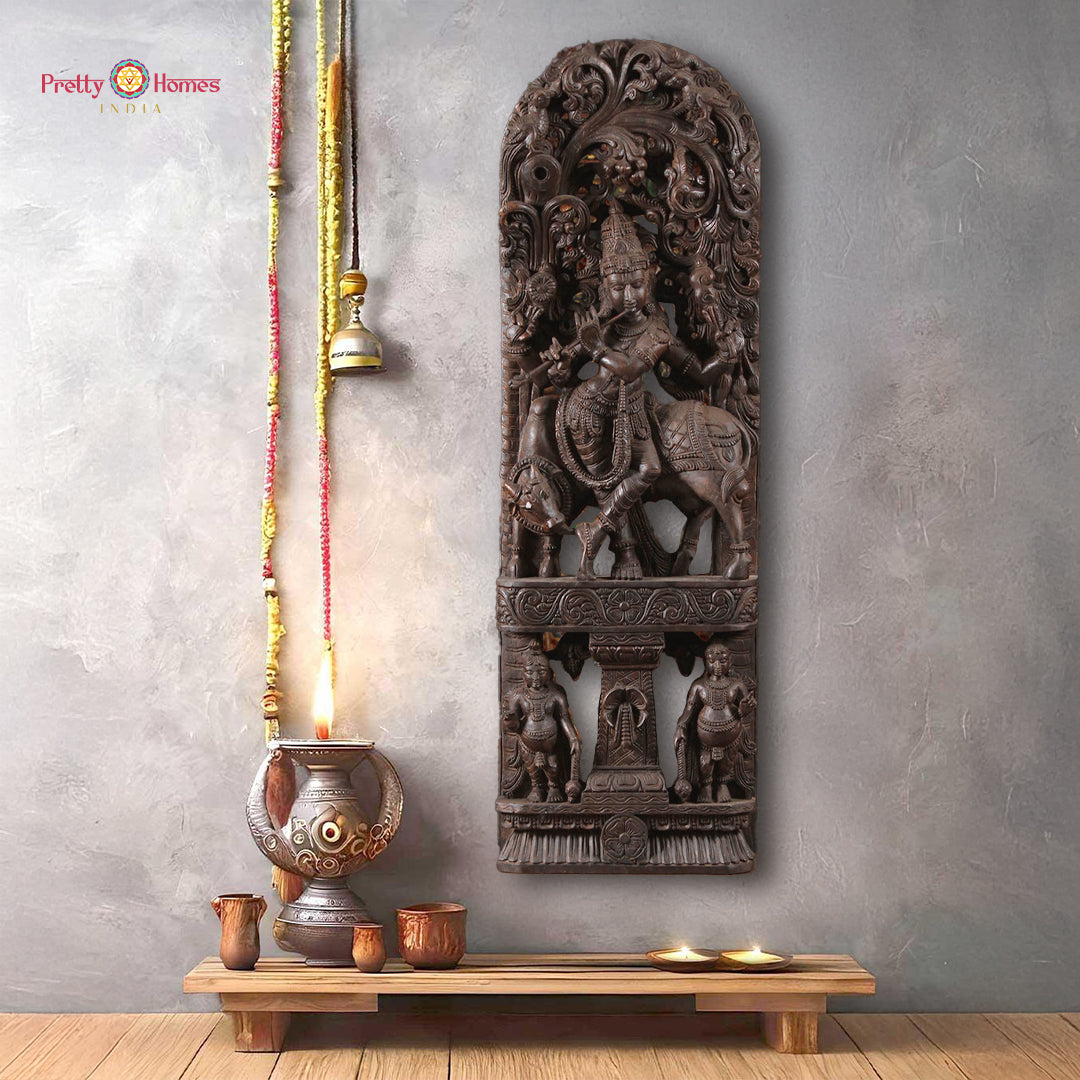 A handcarved wooden sculpture depicting the figure of Krishna playing a flute, accompanied by a cow and two gopis, mounted on a panel canopy.