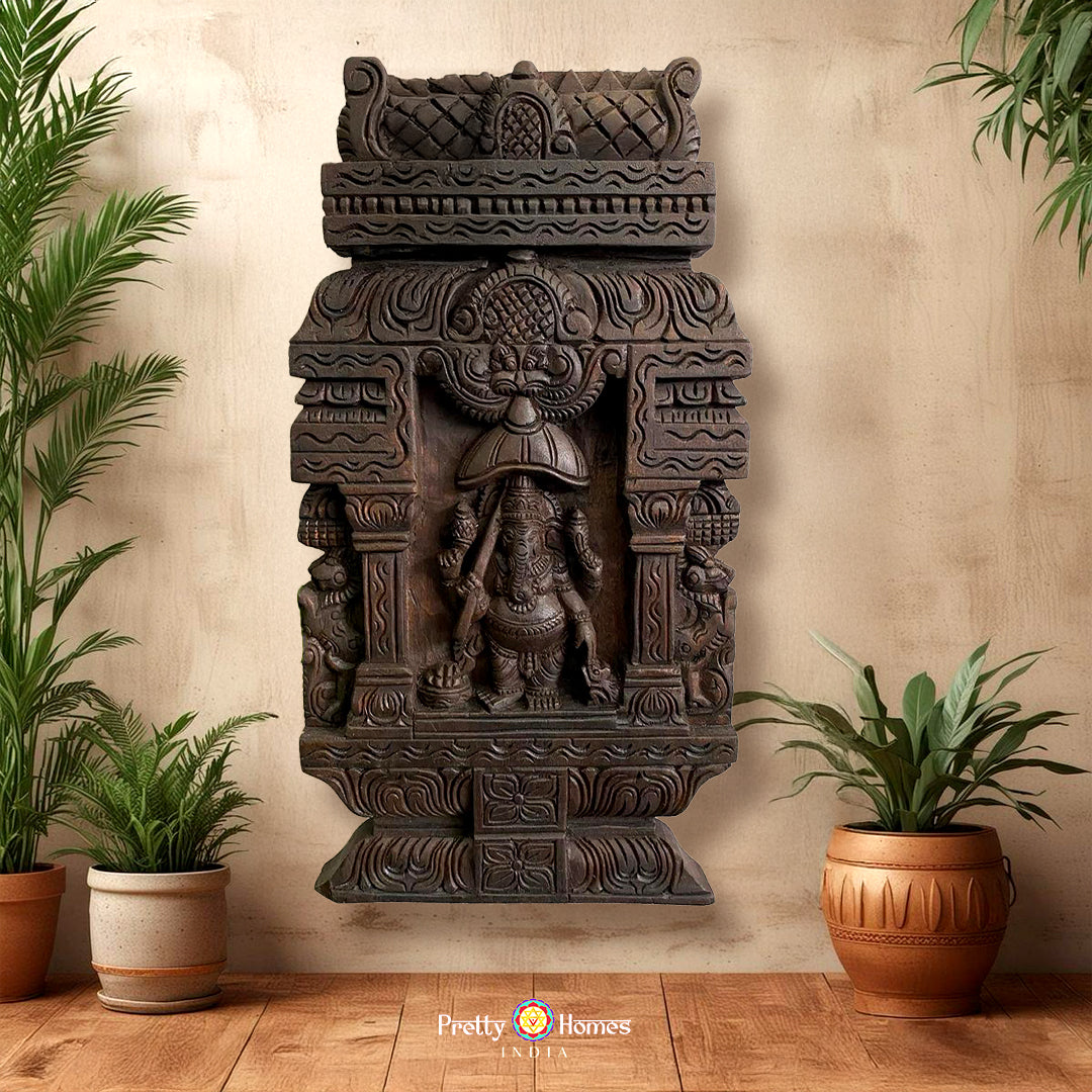 A hand-carved wooden wall panel featuring the figure of Ganesha, placed between two potted plants on a wall.