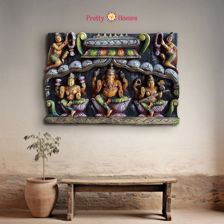 A wooden wall panel featuring carved figures of Ganesh, Laxmi, and Saraswati with Gandharvas on the sides, displayed on a wall. Wooden wall decor panel of Ganesh, Lakshmi, Saraswati with Gandharvas in gopuram design

