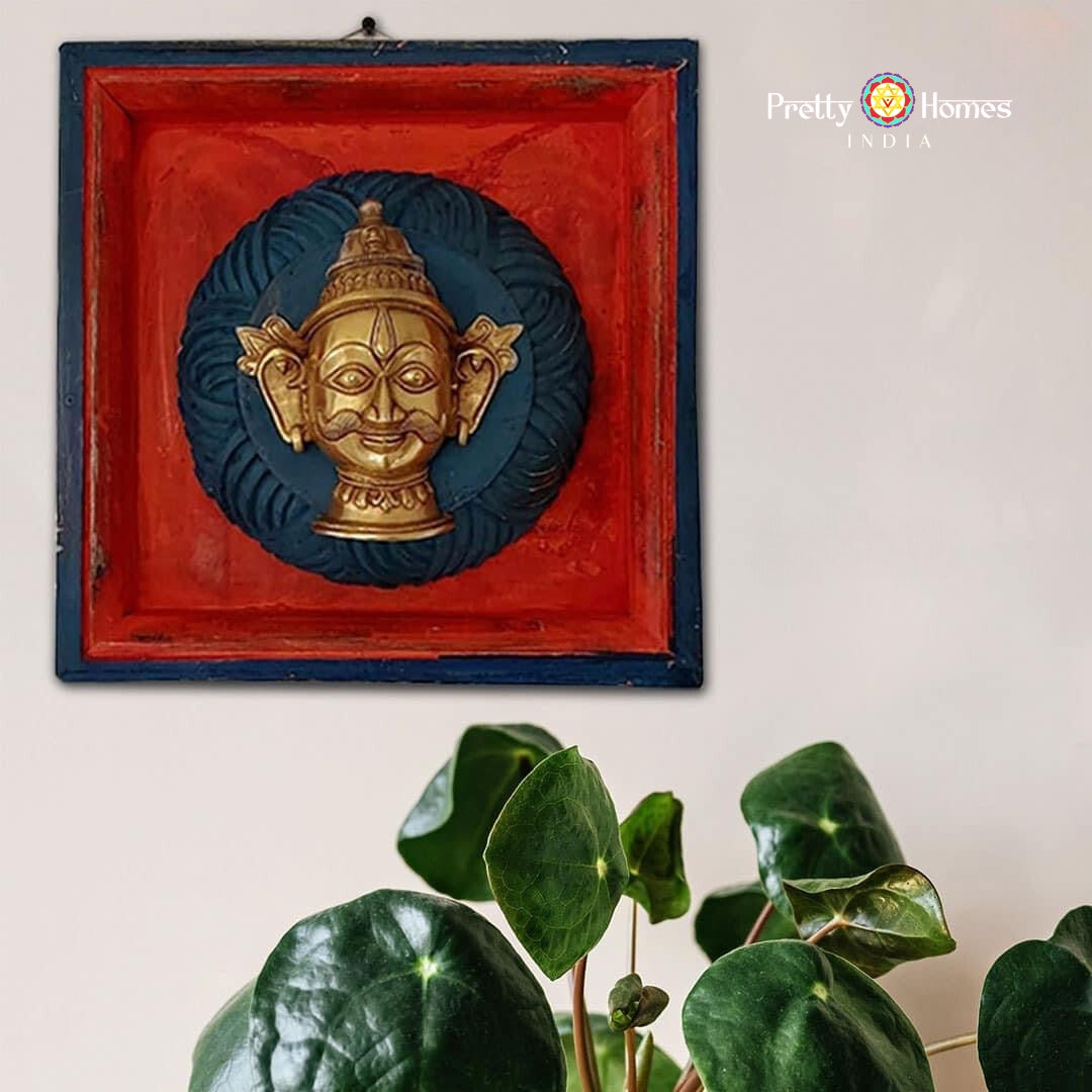 Blue-and-red square frame with brass deity head wall plaque and green plant leaves in foreground.