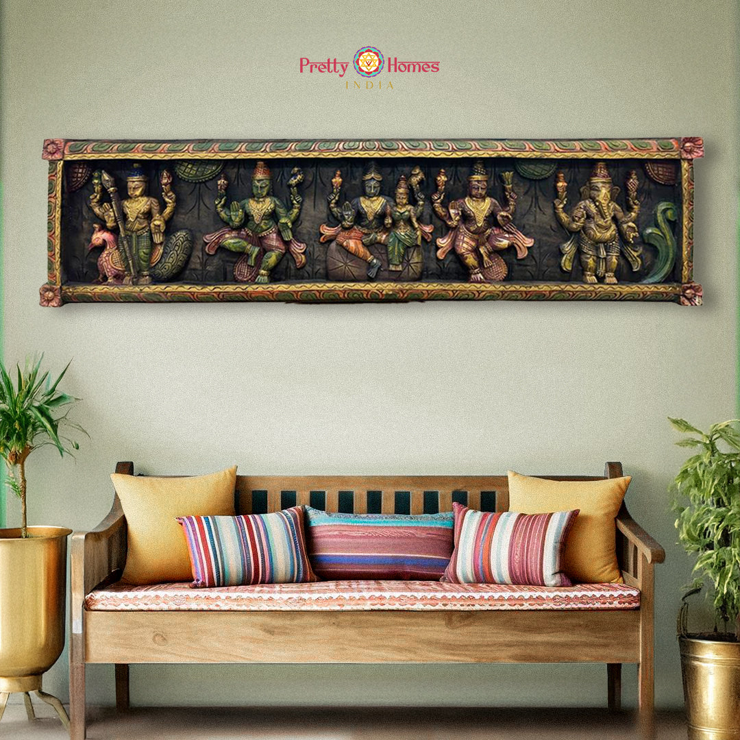 A wall-mounted wooden panel depicting Hindu deities Shiva and Parvati with their family, including Ganesh and Murugan, seated together on a peacock. The panel is colorful and intricately carved, placed above a sofa with cushions in a home setting.