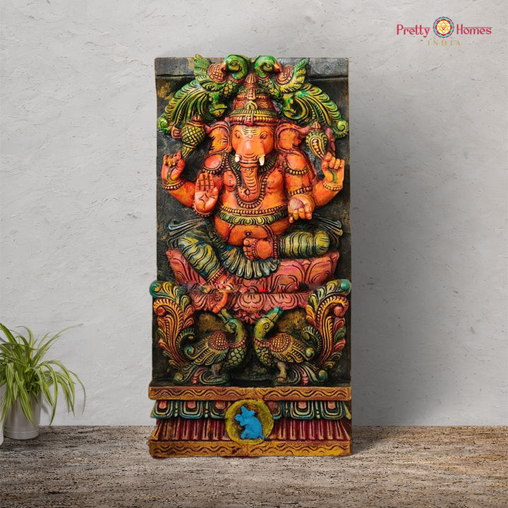 A colorful wooden carved panel featuring an intricately designed figure of Lord Ganesha seated with parrots and peacocks in a vertical orientation.