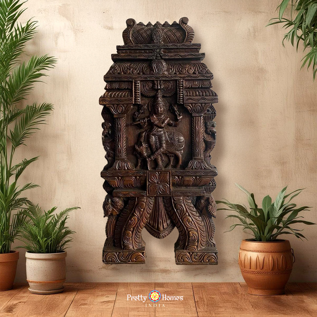 A wall-mounted wooden sculpture of Krishna playing the flute, with intricate carvings and flanked by mythical yalis and parrots, set against a neutral background with potted plants on either side.