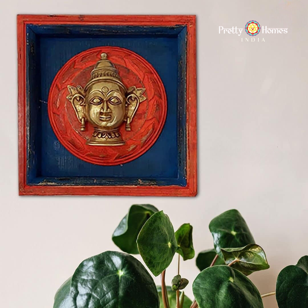 Close-up of blue-framed wall plaque with red circular backing and brass deity-face relief, plant foreground.