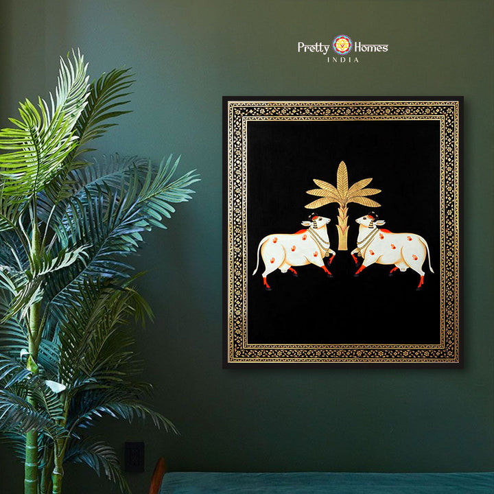 Hand-painted pichwai of two white cows beneath a golden tree on black background, displayed in elegant room.