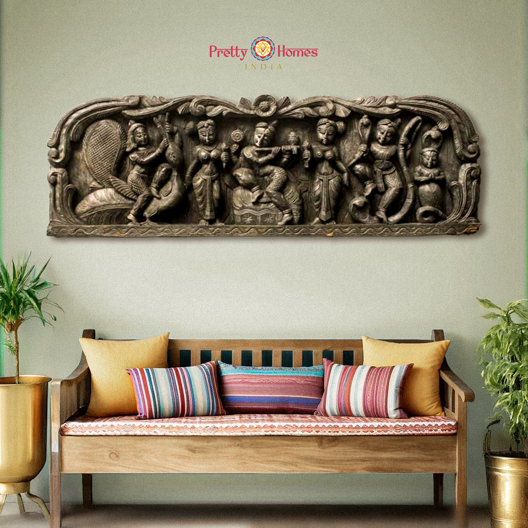 A solid wood wall panel with ornate carvings of Lord Krishna playing the flute, accompanied by cows and Gopikas, mounted on a wall above a bench with cushions.