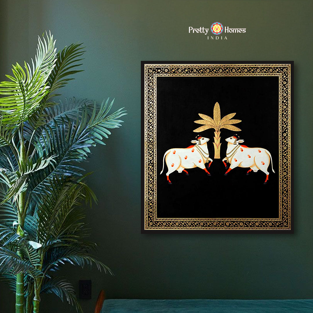 Hand-painted pichwai of two white cows beneath a golden tree on black background, displayed in elegant room.
