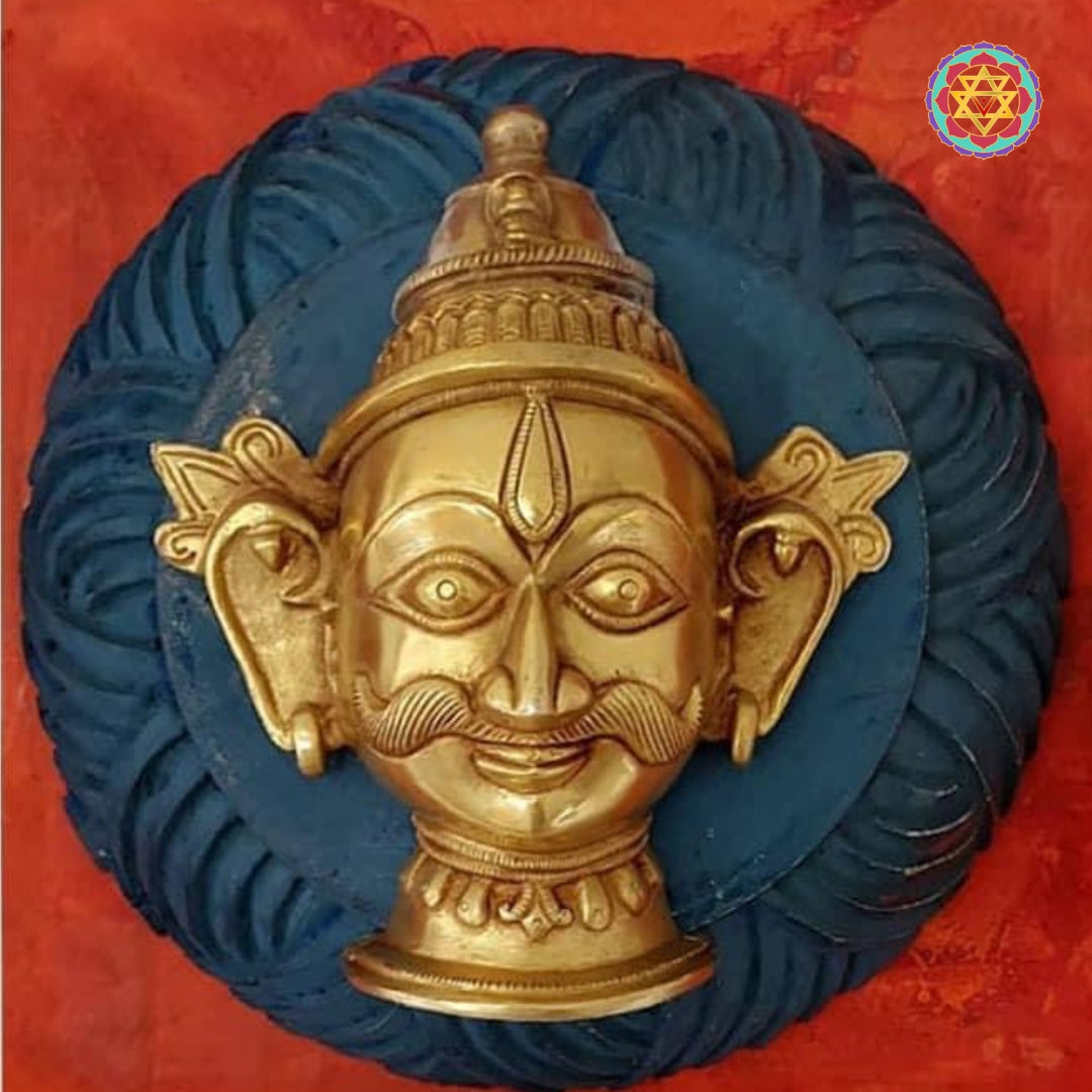 Tight close-up of a small brass deity head relief mounted on a blue circular backing inside a red frame.