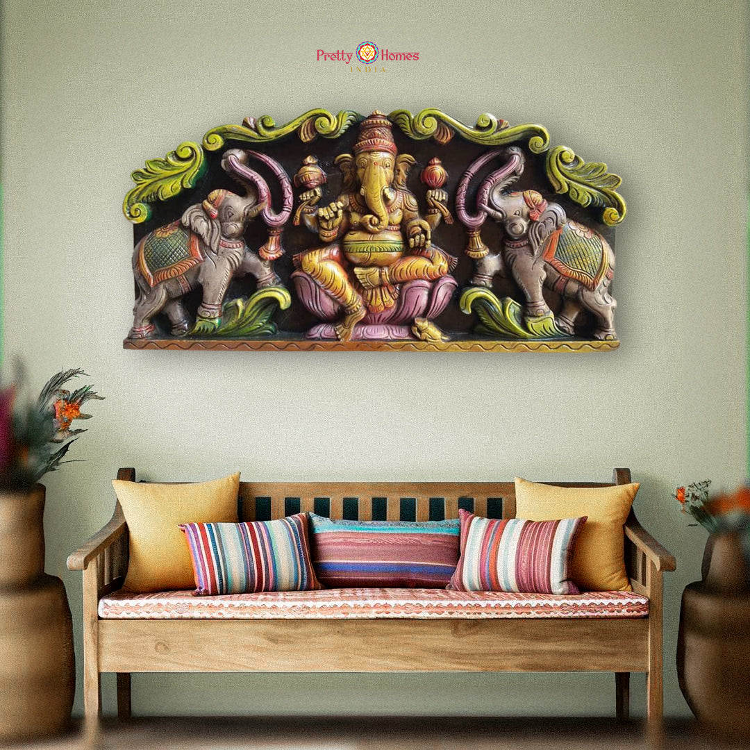 A woodcarved wall panel featuring Lord Ganesha with elephants in vibrant multicolor finish, displayed on a wall above a bench with cushions.