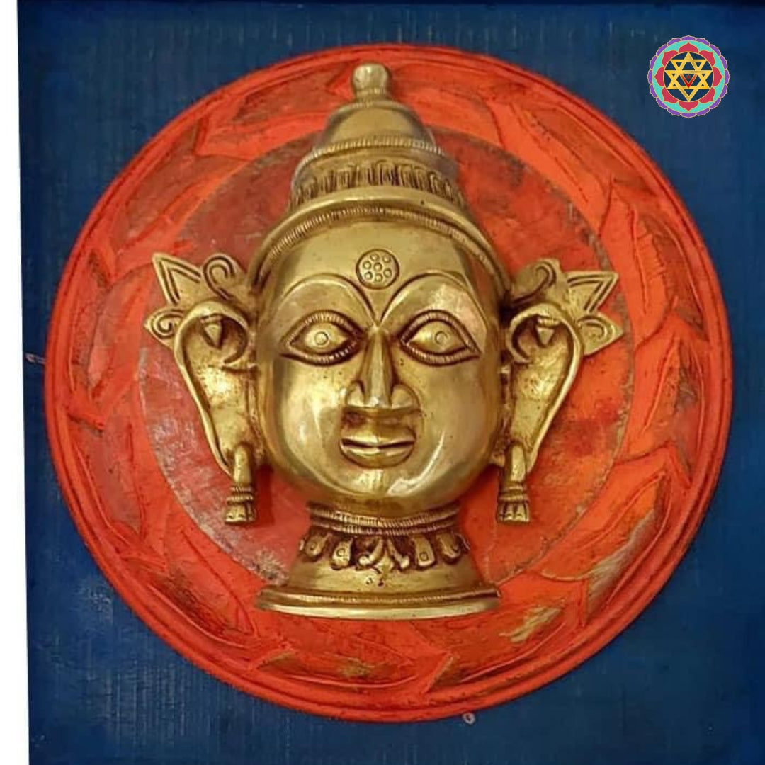 Close-up brass mask with stylized facial features set on a red circular background in a square frame.