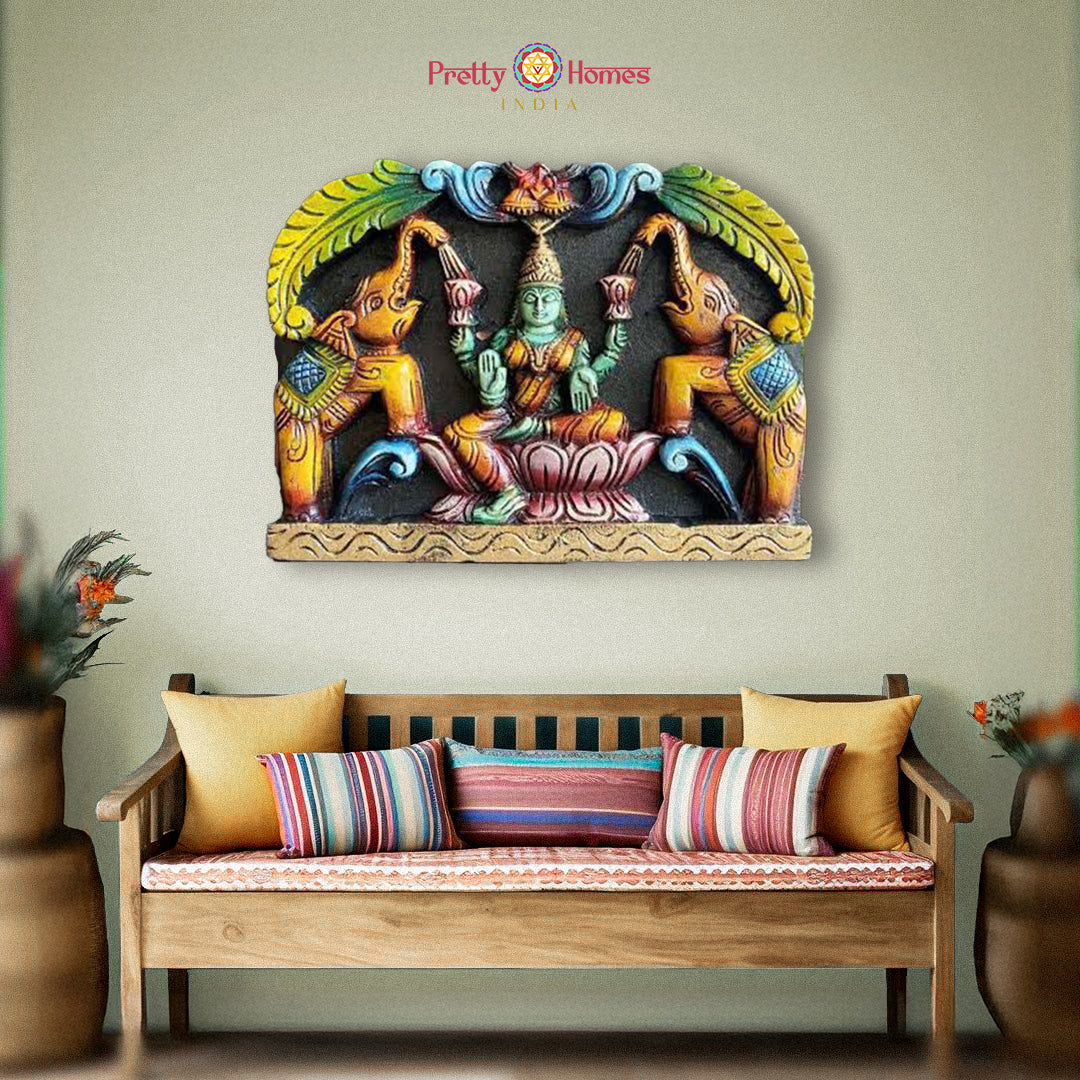 A colorful wall panel featuring Goddess GajaLakshmi with elephants, seated on a lotus, in a protective mudra, displayed above a daybed with cushions.