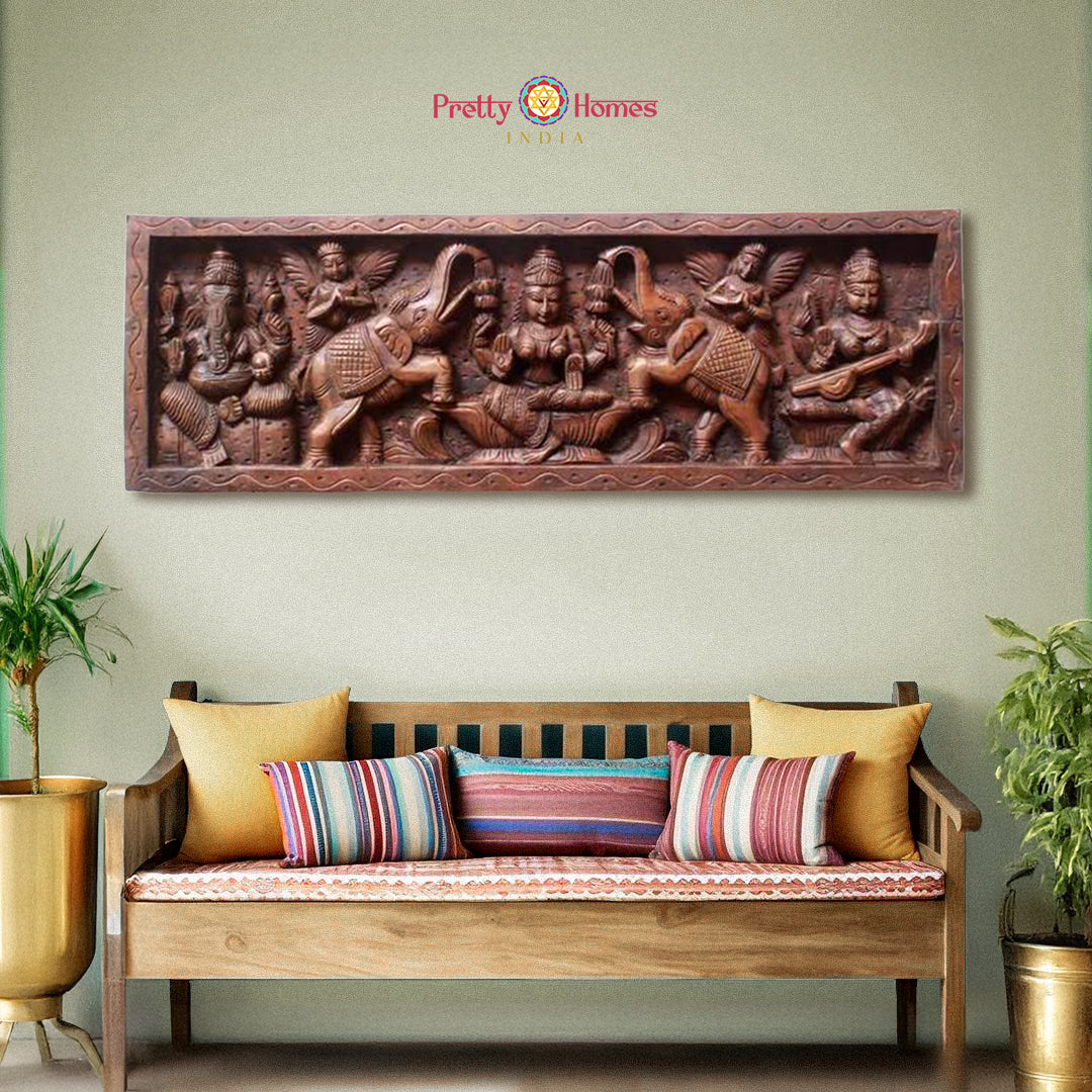 A carved wooden wall panel featuring Hindu deities, with a depiction of Gajalakshmi in the center, Lord Ganesh on one side, and Goddess Saraswati on the other, displayed on a wall above a sofa with cushions.