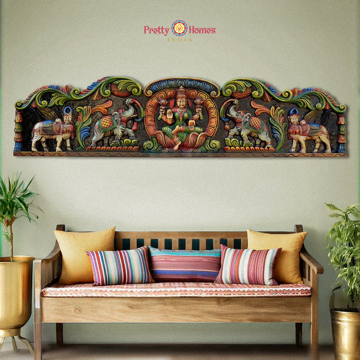 Decorative wall panel with colorful carvings above a wooden bench with cushions. Horizontal wooden wall panels with Gajalaxmi, Kamadhenu, and elephants