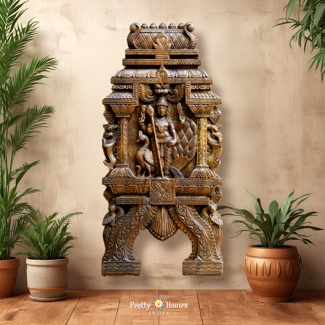 A wall-mounted wooden sculpture of Lord Murugan with a peacock, ornaments, and a flower garland, flanked by mythical yalis and parrots, in a home setting with potted plants on either side.