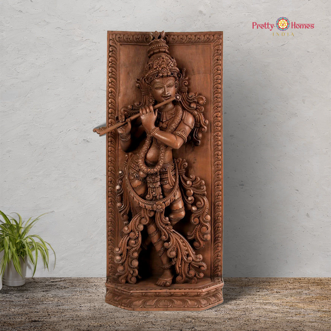 A vertical wooden panel featuring a detailed carving of the Hindu deity Krishna playing a flute, surrounded by floral motifs, with a neutral background.