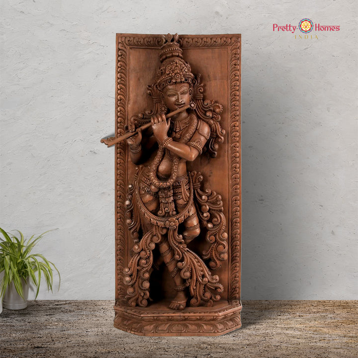 A vertical wooden panel featuring a detailed carving of the Hindu deity Krishna playing a flute, surrounded by floral motifs, with a neutral background.