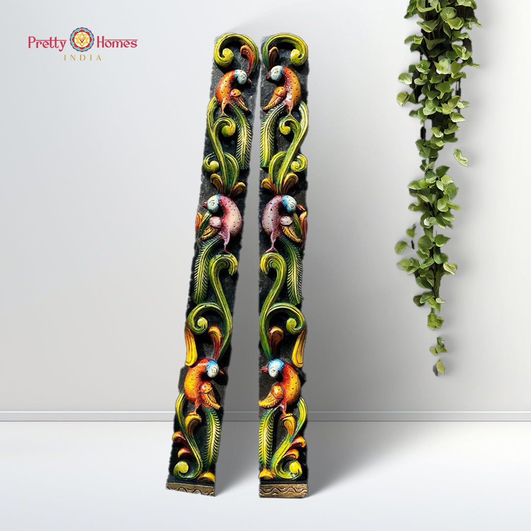 A vertical wall hanging panel with intricate floral, birds, and vine carvings in multicolor design, standing against a white background with a green potted plant beside it.