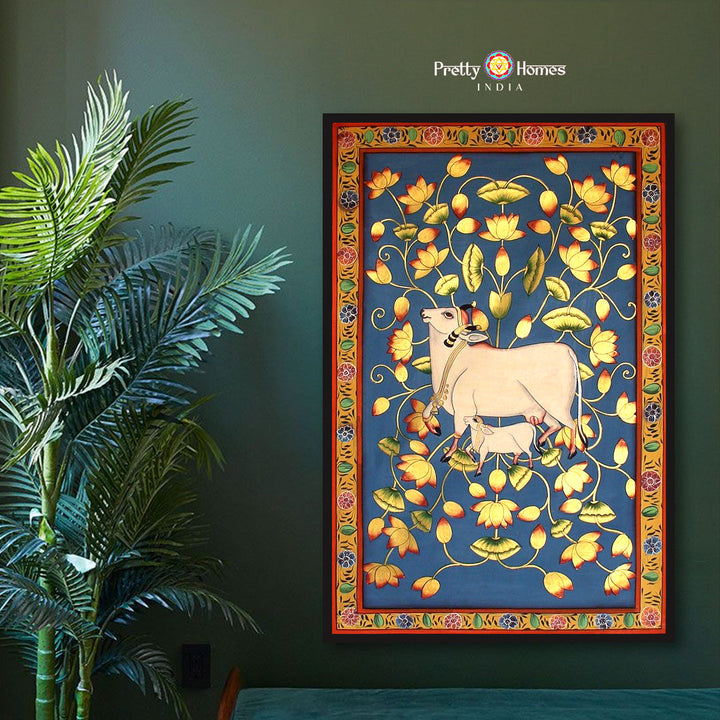 Hand-painted pichwai of a cow and calf standing in a lotus pond, framed in a vibrant red border, displayed on a dark green wall.