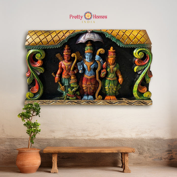 A hand-carved wooden wall panel featuring the divine Hindu deities - Lord Ram, Hanuman, Laxman, Sita with floral carvings, displayed on a wall above a bench with a plant to the side. - Horizontal Ram Darbar panel with Lord Ram, Sita, Laxman & Hanuman

