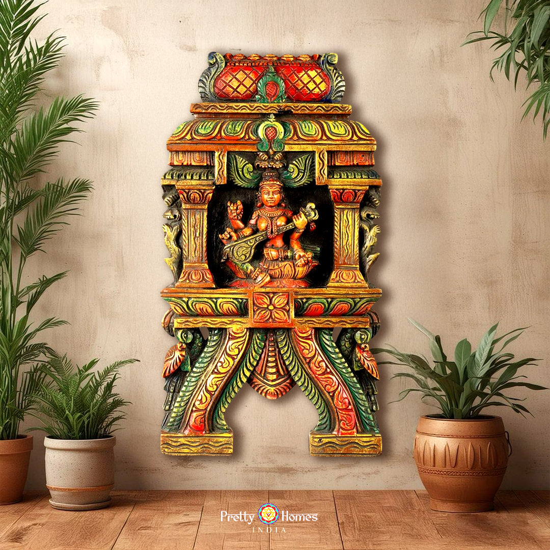 A colorful wall-mounted wooden sculpture of the Hindu goddess Saraswati with peacocks and decorative elements, set against a tropical-themed backdrop.