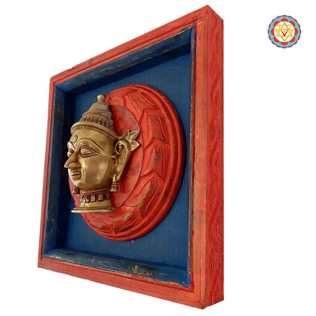 Angled side view of a red square frame with brass deity head mounted on a blue circular backing.